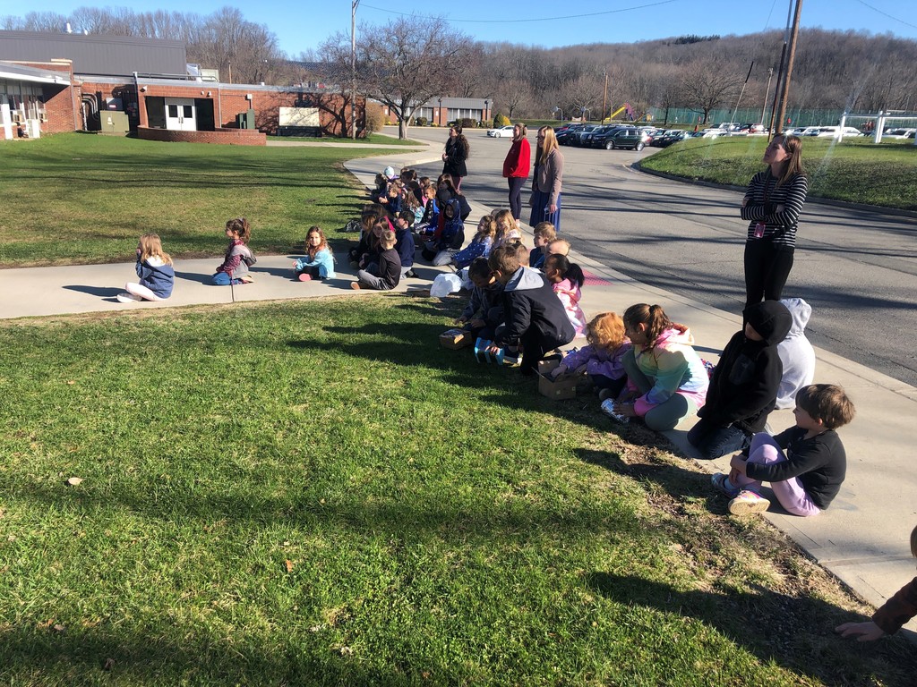 Kindergarten students sit on the sidewalk outside watching their egg drop projects being tested out.